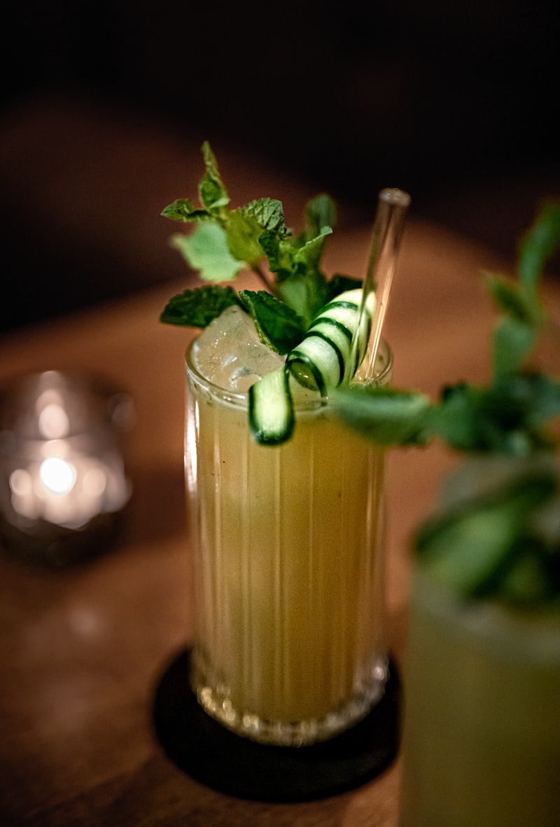 Ein Glas mit gelbem Cocktail, Eis, Gurkenscheiben und Minze dekoriert.